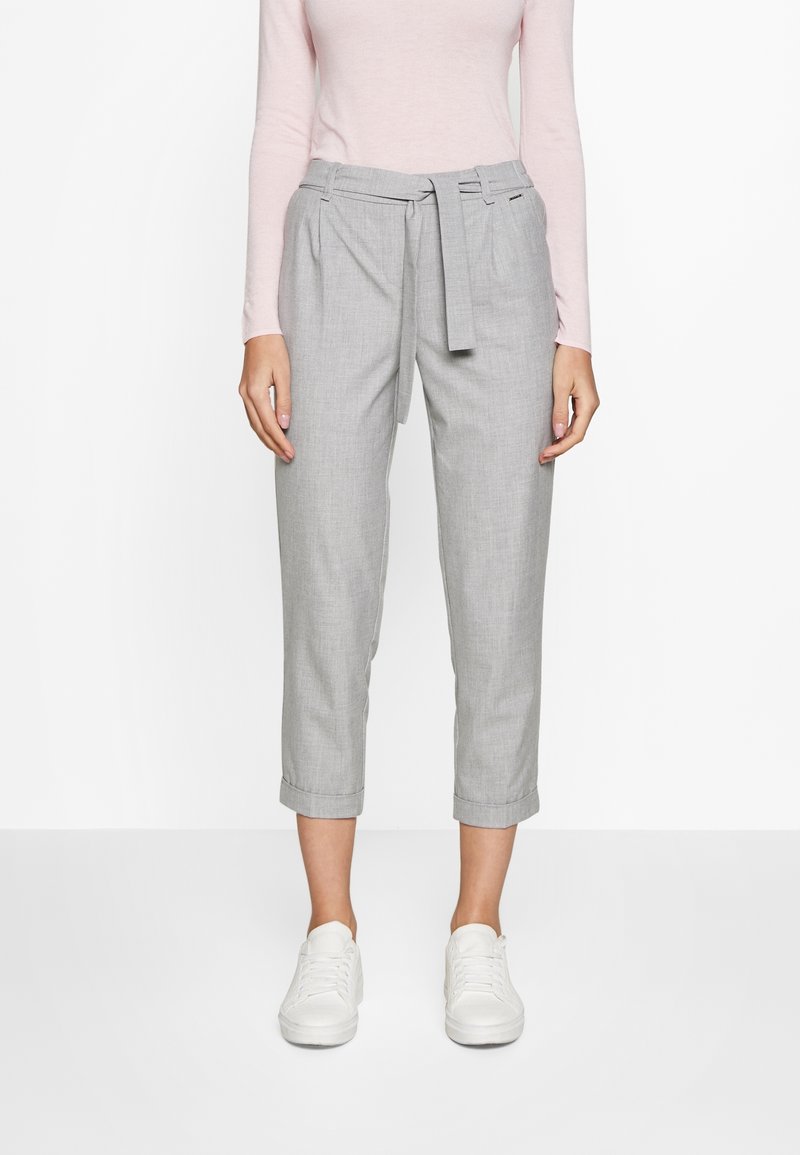 Vrouw in lichtgrijze cropped broek met een stoffen riem, gecombineerd met een lichtroze langemouwen top en witte sneakers.
