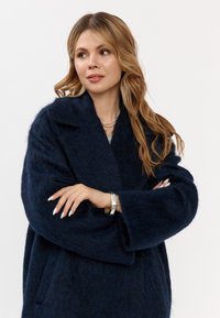 Donna con i lunghi capelli biondi che indossa un cappotto peloso blu scuro, con le braccia incrociate, una collana di perle e un braccialetto d'argento, che guarda di lato.