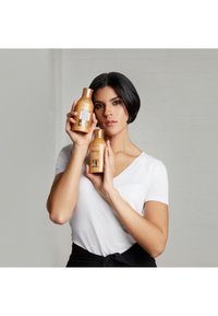 Dwie butelki produktów do włosów Redken, obie w złotym opakowaniu, trzymane przez osobę w białej koszuli, prezentujące etykiety z informacjami o produkcie.