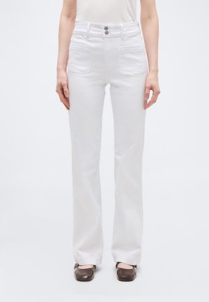 Femme portant un pantalon blanc taille haute à jambes droites avec poches plaquées à l'avant et fermeture à double bouton, associé à des chaussures marron à bout carré.