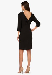 Czarna sukienka z głęboko wyciętymi plecami, rękawami do połowy przedramienia i kokardą przy dekolcie. Gładki materiał i dopasowany krój.