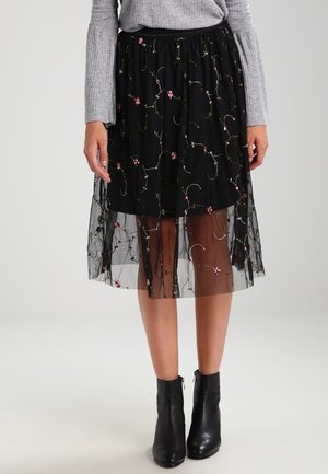 Mujer con falda midi negra con superposición de bordado floral transparente, suéter gris acanalado y botines negros, de pie contra un fondo liso.