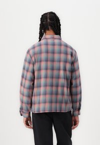 Plaid shirt featuring a blue, pink, and white color scheme, made of soft fabric with a loose fit, and buttoned cuffs. Visible stitching details.