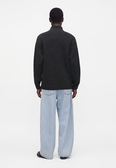 Homme debout de dos, portant un pull tricoté gris foncé, un jean large bleu clair et des chaussures noires sur fond blanc.