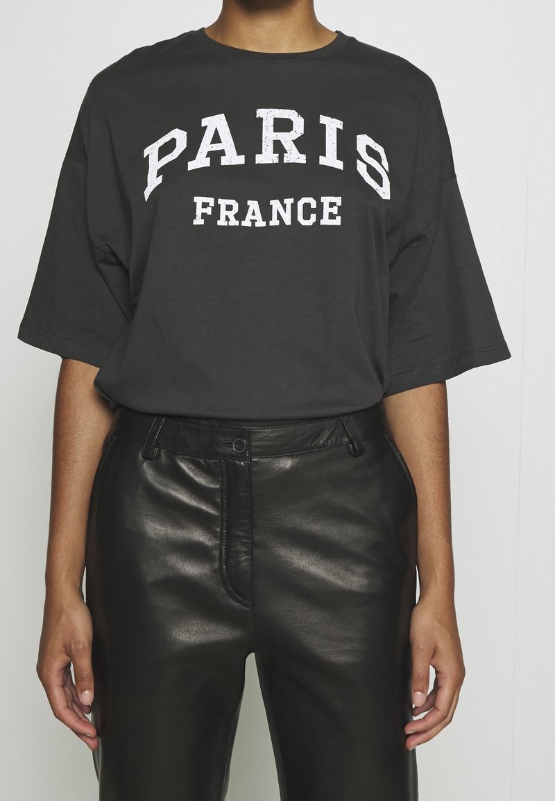 Graues, übergroßes T-Shirt mit "PARIS" und "FRANCE" in Weiß bedruckt. Kombiniert mit hochgeschnittenen, schwarzen Lederhosen, die eine glatte Textur zeigen.