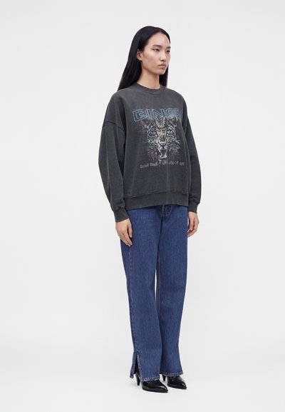 Femme portant un pull oversize gris foncé avec un motif visage de léopard et un jean bleu à jambes larges avec des fentes à l'avant et des bottes noires à talons.