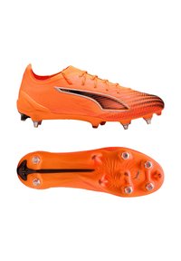 Narancssárga focicipő texturált felsőrésszel, fekete díszítésekkel és masszív talppal, amelyen hat kúpos stopli található a tapadás érdekében.