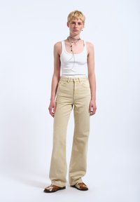 Jong persoon met kort blond haar, die een wit tanktopje, beige broek met hoge taille, gelaagde kettingen en bruine schoenen draagt.