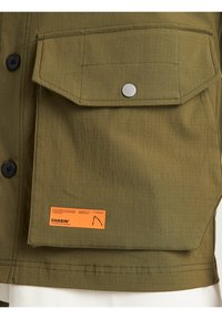 Zbliżenie na kieszeń oliwkowej kurtki z klapą i srebrnym guzikiem, pomarańczowa metka z czarnym tekstem oraz czarne guziki na przedniej części kurtki.