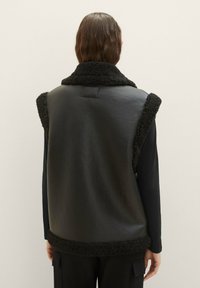Schwarze Weste mit Kunstfellbesatz, ärmelloses Design, glattes Leder-Material und hohem Kragen. Einfache Naht auf der Rückseite.