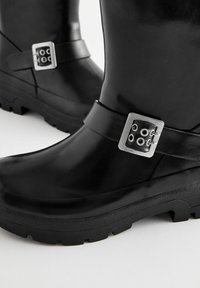Des bottes de cheville noires en caoutchouc présentent une semelle texturée, une large sangle avec une boucle en métal argenté et une finition lisse et brillante.