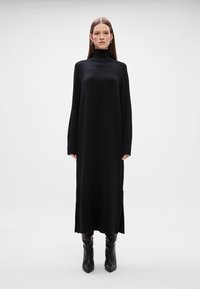 Μαύρο πλεκτό φόρεμα με ψηλό λαιμό, μακριά μανίκια, πλαϊνές σχισμές και άνετη εφαρμογή, που συνδυάζεται με μαύρες μπότες μέχρι το γόνατο.