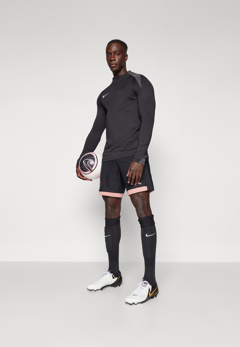Nike Sportbekleidungsset, schwarzes Langarmoberteil, Shorts mit pinkfarbenem Saum, schwarze kniehohe Socken, weiße Schuhe mit schwarzen Akzenten, hält einen Fußball.