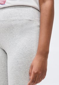 Κοντινό πλάνο ενός ατόμου που φοράει ανοιχτό γκρι φόρμα με εφαρμοστή μέση, δείχνοντας το δεξί του χέρι να ακουμπά δίπλα στο ισχίο.