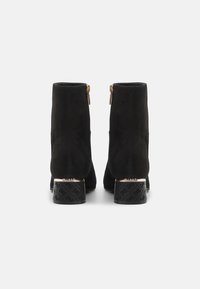 Stivaletti alla caviglia in suede neri con punta arrotondata, dettaglio della cerniera dorata e tacco a blocco decorato con una base nera e accenti metallici.