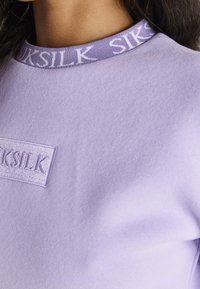 Fialově lila mikina s texturovaným povrchem, vybavená žebrovaným výstřihem a vyšívanou logovou náplastí. Design je jednoduchý a čistý.