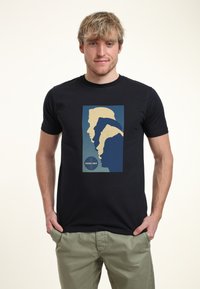 Svart T-shirt med en stiliserad grafik av profiler i nyanser av blått och beige, med "PAN AM"-logotypen i botten. Mjukt bomullstyg.