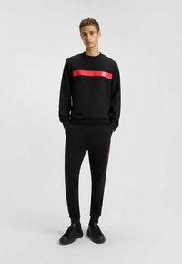 Set di felpa e pantaloni neri, con una striscia orizzontale rossa e logo. Tessuto liscio, polsini elasticizzati e design slim fit. Sneakers nere.