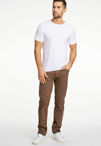 Witte t-shirt met korte mouwen, gecombineerd met een bruine broek en witte sneakers. Model staat casual met de hand op de heup tegen een effen achtergrond.