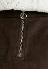 Hnědá semišová látka se stříbrným zipem a kulatým táhlem. Textura je hladká, zdůrazňující elegantní design oděvu.