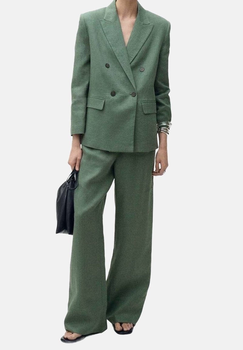 Personne portant un blazer vert ample à double boutonnage et un pantalon large assorti, tenant un sac à main noir, avec des bracelets argentés.