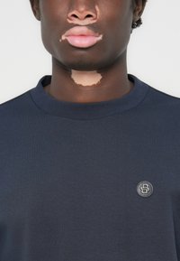 Marineblå kortermet genser med rund hals, som har et teksturert logomerke på venstre bryst og en glatt stofffinish.