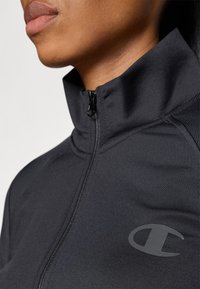 Černá sportovní bunda s vysokým límcem a zipem na přední straně. Obsahuje decentní logo na hrudi a hladkou, přiléhavou texturu.