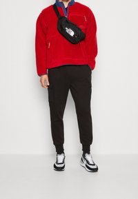 Chaqueta de forro polar roja con collar con cremallera, pantalones cargo negros con bolsillos laterales y zapatillas en blanco y negro. Bolsa cruzada ajustable negra.