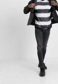 Maglione a righe bianche e nere, giacca nera, jeans neri strappati e sneakers nere, che mostrano un look casual a strati.