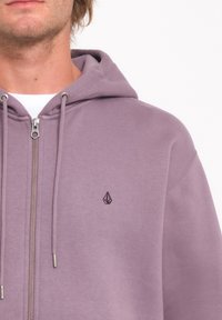 Sudadera con cremallera morada, fabricada en un tejido suave, que cuenta con una capucha con cordón y un pequeño logo negro en el lado izquierdo del pecho. Detalle de cremallera plateada presente.