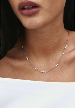 Gros plan d’une personne portant un délicat collier en chaîne argentée avec de petites perles blanches, associé à un haut blanc épaules dénudées.
