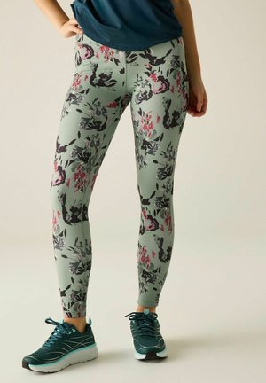 Person trägt Leggings mit Blumenmuster und türkisfarbene Sportschuhe und steht mit einer Hand in der Hüfte vor einfachem Hintergrund.