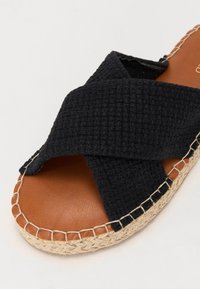 Sorte vevd tekstil sandal med krossede stropper, brun skinnfot seng, og naturlig tau såledetalj. Rund tå og sydde detaljer.