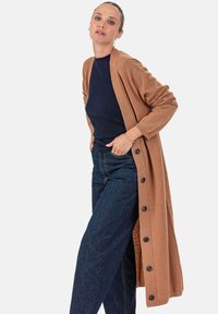 Langer brauner Cardigan mit weicher Textur, vorne mit Knöpfen und offenen Seiten, darüber getragen ein navyblauer Pullover und dunkle Jeans.