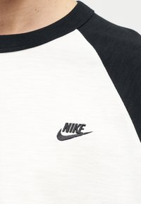 Κοντινό πλάνο ενός λευκού πουκαμίσου με μαύρα raglan μανίκια που διαθέτει ένα μικρό μαύρο λογότυπο Nike στο στήθος.