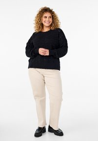 Pull en tricot noir avec un motif texturé, col rond et poignets côtelés, associé à un pantalon beige et des chaussures noires chunky.