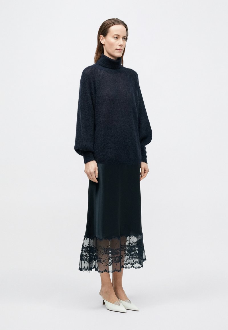 Dark knitted turtleneck sweater paired with a black silky skirt featuring a lace hem. The outfit is complemented by white, pointed-toe shoes.