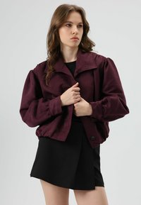 Bordeauxrode jas met opgeblazen mouwen, knoopsluiting en een kraag. Gedragen over een zwarte rok. Zachte textuur, casual ontwerp.