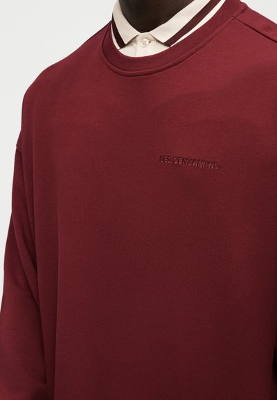 Άνδρας φορώντας μπορντό φούτερ με το κέντημα «LES BENJAMINS» στο στήθος, από πάνω από ένα εκρού πουκάμισο με καφέ ρίγα στο γιακά.