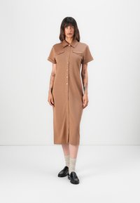 Brun knappete kjole med kort erm, to brystlommer og rett snitt. Brukt med svarte loafers og beige knelange sokker.