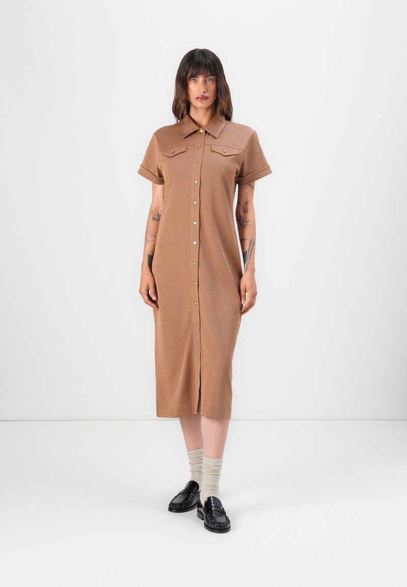 Brun knappete kjole med kort erm, to brystlommer og rett snitt. Brukt med svarte loafers og beige knelange sokker.