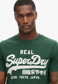 Camiseta de algodón verde con un gran logo impreso en blanco que incluye el texto "REAL SuperDry" y caracteres japoneses adicionales. Mangas cortas, cuello redondo.