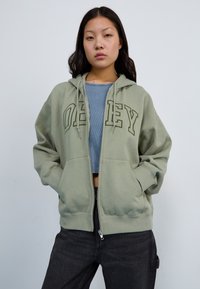 Green zip-up hoodie with "OBEY" text, soft fabric, large front pockets, and a drawstring hood. Worn over a light blue top and dark jeans.