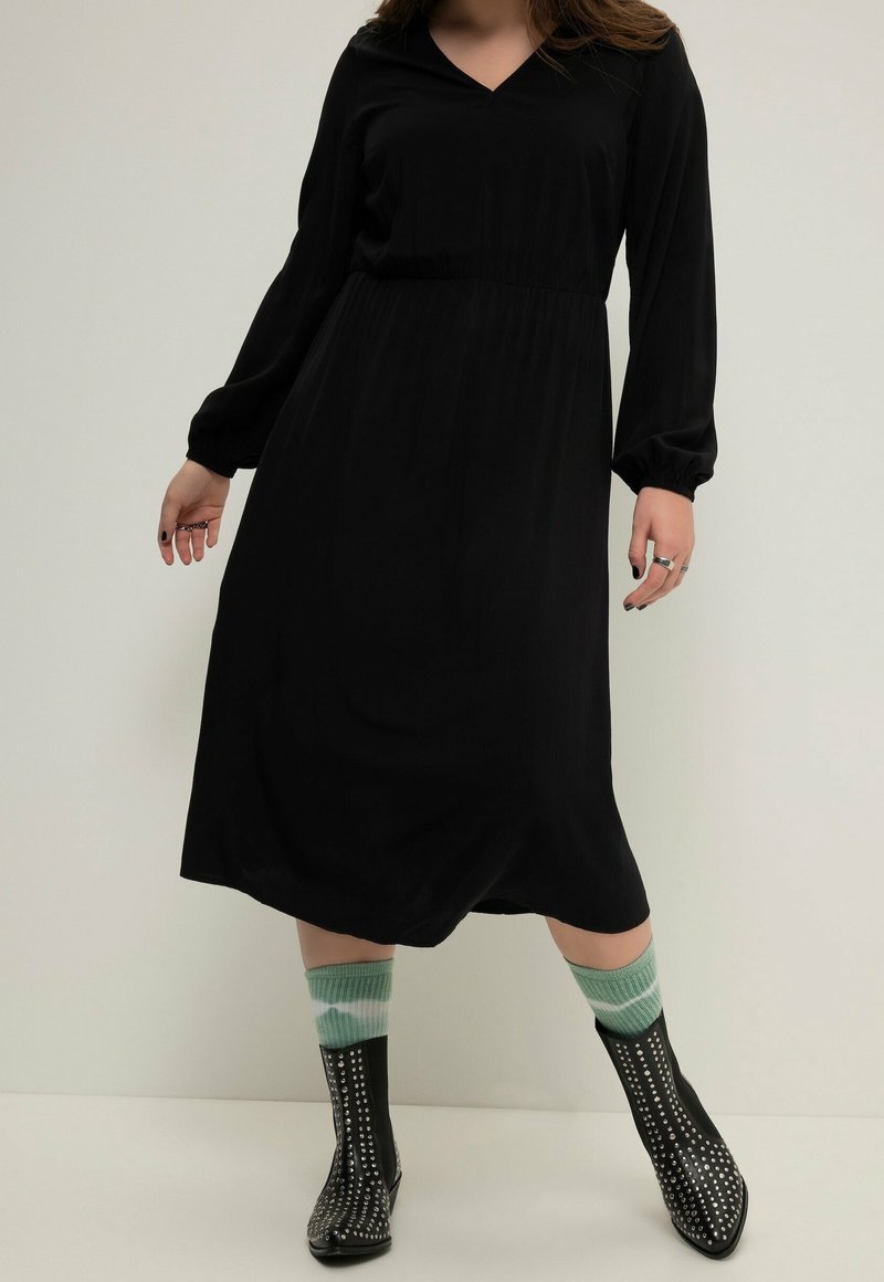 Personne portant une longue robe noire, des chaussettes vertes rayées et des bottines noires cloutées, debout devant un fond uni.