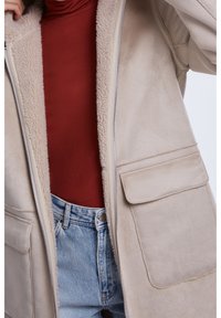 Rozepnutá béžová kožešinová bunda přes hnědou rolákovou košili a světle modré džíny.