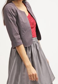 Femme portant une veste courte grise sur un haut rouge brodé et une jupe en satin grise taille haute sur fond blanc.