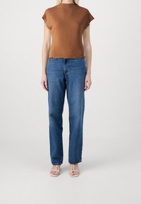 Kvinde iført en kortærmet, brun tekstureret top, lige ben blå jeans og hvide, stroppede sandaler med åben tå på en ensfarvet lys baggrund.