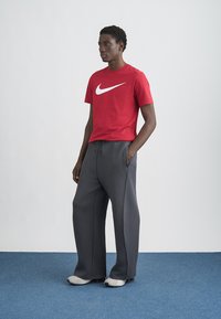 Κόκκινο κοντομάνικο μπλουζάκι Nike με λευκό λογότυπο, σε συνδυασμό με γκρι φαρδιά αθλητική παντελόνα. Το μοντέλο στέκεται σε μπλε χαλί μπροστά σε απλό φόντο.