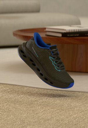 Chaussure de sport Skechers noire et bleue avec semelle coussinée flottant au-dessus d'un tapis beige dans un salon avec une table ronde en bois et des livres.