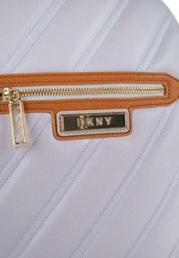 Bolso de cuero púrpura claro con costuras diagonales, detalles de cuero marrón, herrajes dorados y una placa con el logo de DKNY cerca de un bolsillo con cremallera.